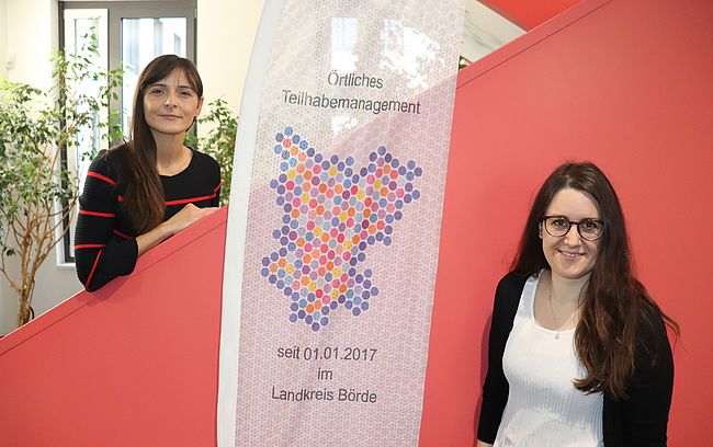 Das Foto zeigt die Innentreppe des Landratsamtes in Haldensleben, davor ein Banner mit der Aufschrift "Örtliches Teiilhabemanagement" und Katja Klommhaus (links) und Anne Sophie Fischer als direkte Ansprechpersonen für Angelegenheiten von Menschen mit Beeinträchtigungen. 