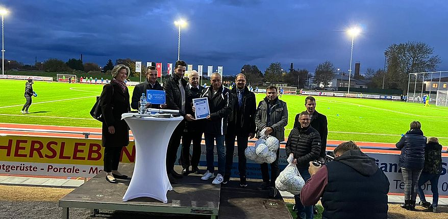 Das Foto zeigt einen Blick in das Stadion. Im Vordergrund offizielle Akteure des Tages, unter anderem Landrat Martin Stichnoth, Bürgermeister Benjamin Kanngießer und Joachim Kunkel, Vorsitzender des OSC. Im Hintergrund sieht man den vom Flutlich bestrahlten neuen Kunstrasenplatz.