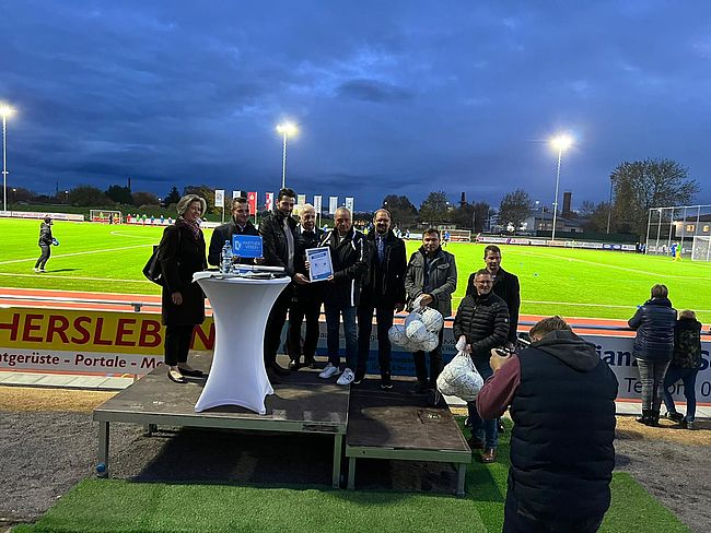 Das Foto zeigt einen Blick in das Stadion. Im Vordergrund offizielle Akteure des Tages, unter anderem Landrat Martin Stichnoth, Bürgermeister Benjamin Kanngießer und Joachim Kunkel, Vorsitzender des OSC. Im Hintergrund sieht man den vom Flutlich bestrahlten neuen Kunstrasenplatz.