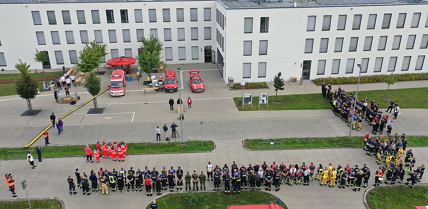Die Luftbildaufnahme ist am 11. September 2021 zum Übungsbeginn mit einer Drohne am Verwaltungsgebäude des Landkreises in Haldensleben entstanden. Es zeigt die an der Übung beteiligten Einheiten, Menschen und Technik, aus der Vogelperspektive. Im Vordergrund haben die Einheiten Aufstellung auf dem Parkplatz genommen. Im Hintergrund ist das Verwaltungsgebäude "Bornsche Straße 2" zu sehen.