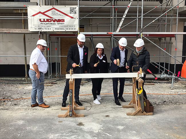 Das Foto zeigt im Hintergrund einen kleinen Ausschnitt des Rohbaugebäudes. Davor ein Holzbock, in den Ralf Kürbis und Sylvia Franke (beide DRK), die Pflegedienstleiterin des Wohnquartiers Oebisfelde, Katja Meinke, und der Landrat die letzten Nägel vor dem Richtfest eintreiben. Beobachter des Geschehens ist Hans-Werner Kraul, Bürgermeister von Oebisfelde-Weferlingen.   