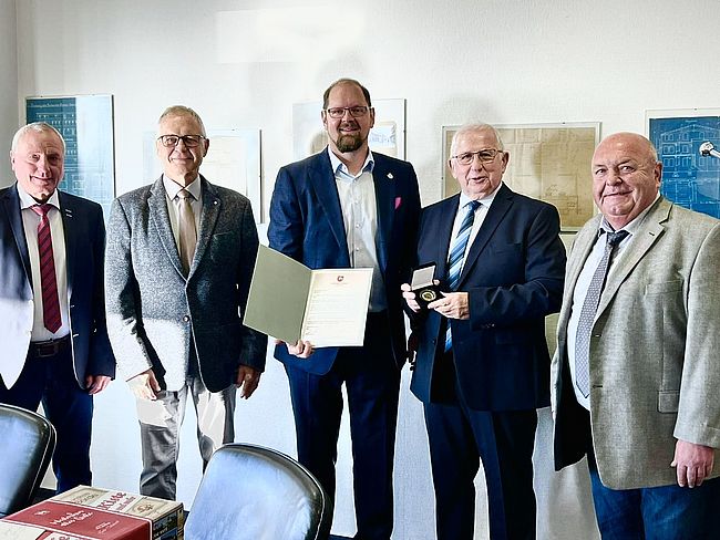 Das Foto zeigt Landrat Martin Stichnoth und Joachim Kunkel mit Urkunde und Erinnerungsmedaille. Daneben 3 Herren des Unternehmens und des Oscherslebener Sportclubs. Das Foto ist aufgenommen in einer Räumlichkeit der "oddesse". 