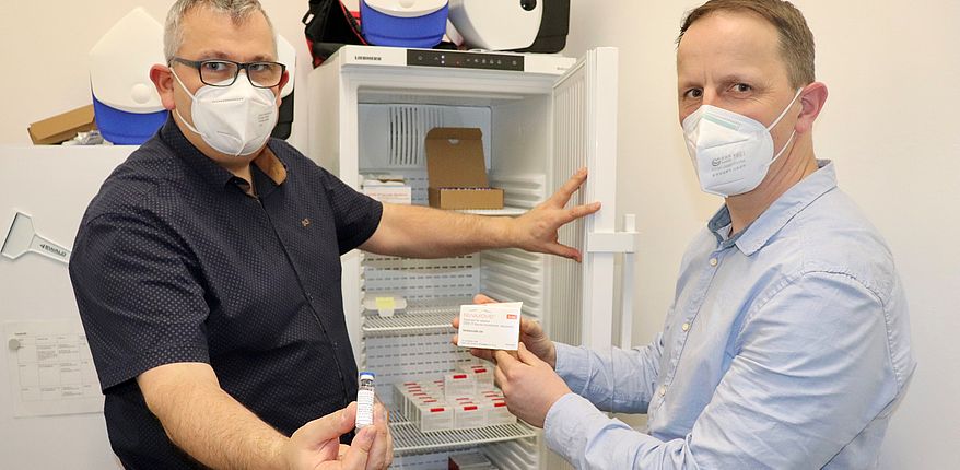 Mario Bernsdorf und Stefan Crackau (Impfstelle Landkreis Börde) zeigen vor einem geöffneten Kühlschrank die Impfdosen von Novavax 