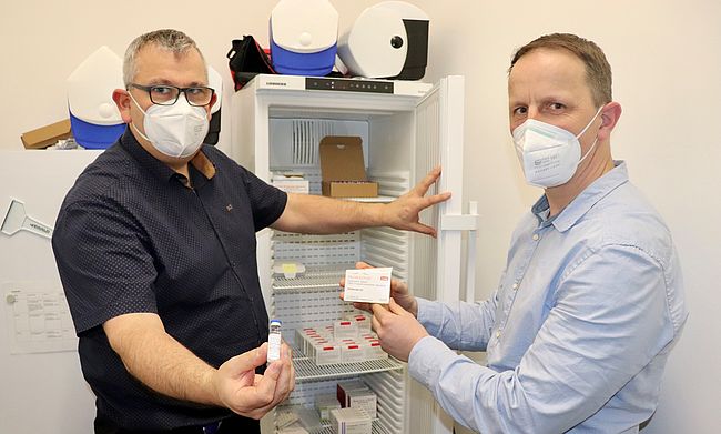 Mario Bernsdorf und Stefan Crackau (Impfstelle Landkreis Börde) zeigen vor einem geöffneten Kühlschrank die Impfdosen von Novavax 