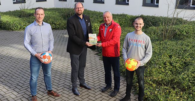 Das Foto zeigt Dietrich Lehmann bei der Übergabe des Buches an Martin Stichnoth, daneben zwei sportbegeisterte Mitarbeiter der Kreisverwaltung, links Domenik Siegmund, selbst aktiv beim Eilslebener Sportverein, und Kilian Paetzel, Nachwuchstrainer „NFZ“ Oschersleben, ausgestattet mit der C-Trainer-Lizenz.