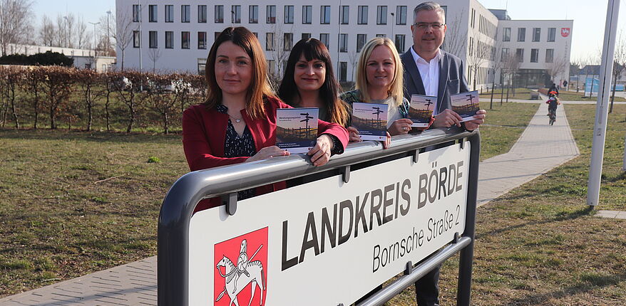 Stefanie Kunz, Netzwerkkoordinatorin, Familienhebamme und Koordinatorin Christina Stolarczyk präsentieren den Beratungswegweiser. Sie stehen hinter dem Einfahrtsschild mit der Aufschrift "Landkreis Börde" zum Parkplatz des Verwaltungsgebäudes in Haldensleben.