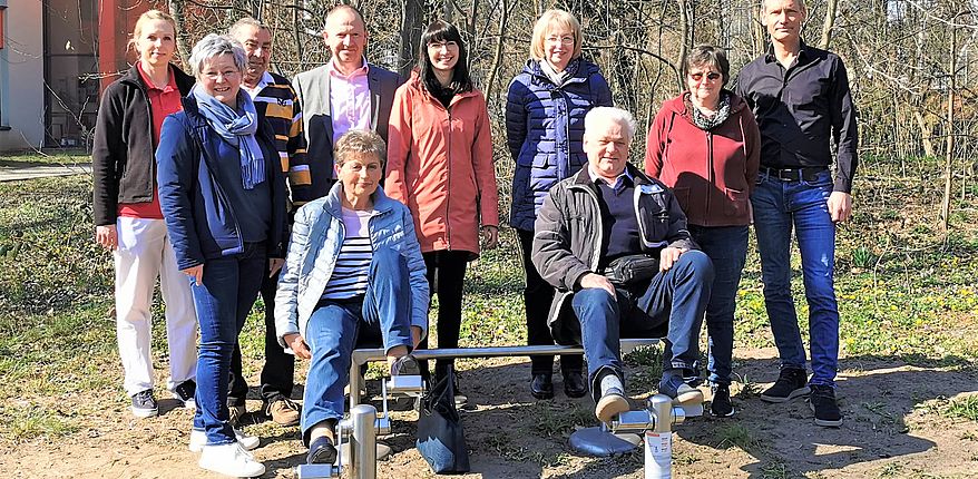 Foto Stadt Haldensleben / Sabine Wendler, Rüdiger Mages und Dr. Elena Sterdt freuen sich mit den Gästen über die Einweihung des Bewegungs-Parcours in Althaldensleben. Im Vordergung wird die Beinpresse getestet. Im Hintergrund stehen die gäste der Einweihung zum Erinnerungsfoto. 