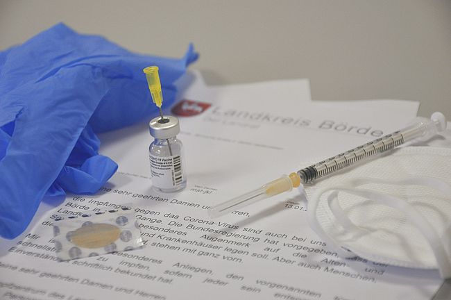 Das Foto zeigt ein Impffläschchen, eine Spritze und einen schriftlichen Bescheid des Landkreises Börde auf einem Kopfbogen.