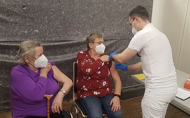 Das Foto zeigt einen impfberechtigte Person bei der Impfung. Eine Person wird gerade geimpft, eine zweite Person wartet. 