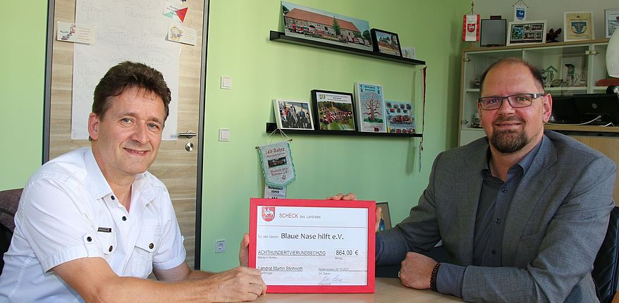 Das Foto zeigt Landrat Martin Stichnoth und Roger Altenburg an einem Beratungstisch im Landratsamt in Haldensleben. Beide halten einen symbolischen Scheck über 864 Euro. Altenburg ist Vorsitzender des Vereins "Blaue Nase hilft".