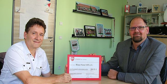 Das Foto zeigt Landrat Martin Stichnoth und Roger Altenburg an einem Beratungstisch im Landratsamt in Haldensleben. Beide halten einen symbolischen Scheck über 864 Euro. Altenburg ist Vorsitzender des Vereins "Blaue Nase hilft".